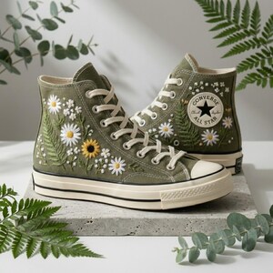 Puede incluir: Zapatillas altas verde oliva con margaritas blancas bordadas, helechos y un girasol. Las zapatillas tienen cordones blancos, puntera color crema y el logo Converse All Star en el lateral.