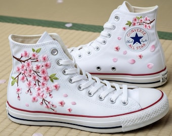 May include: White high-top sneakers with embroidered pink cherry blossoms and green leaves. The sneakers have a red and blue stripe on the sole and the Converse All Star logo.