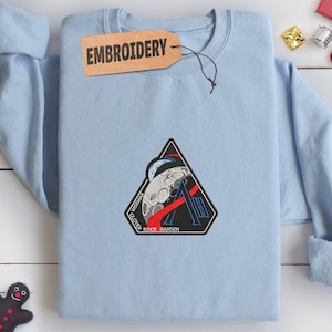 以下が含まれることがあります： 月とWiseman、Glover、Koch、Hansenの名前が刺繍された、黒、赤、白のパッチが付いた、水色のクルーネックのスウェットシャツ。「EMBROIDERY」という文字が入った茶色のタグがネックラインからぶら下がっています。