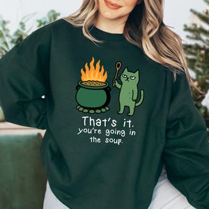 May include: Dark green sweatshirt with a humorous graphic. The design features a green cat holding a wooden spoon, a bubbling cauldron with flames, and the text "That's it. You're going in the soup."