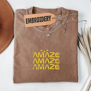 Könnte beinhalten: Ein braunes T-Shirt mit dem Wort "EMBROIDERY" auf einem Etikett. Das Shirt hat das Wort "AMAZE" in Gelb gestickt, mit einer silbernen Halskette und einem braunen Hut im Hintergrund. Das Shirt ist gefaltet.