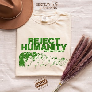Puede incluir: Camiseta color crema con el texto "REJECT HUMANITY RETURN TO FROG" en verde. La camiseta presenta un gráfico de rostros humanos que se transforman en ranas. Un sombrero marrón y flores secas están en el fondo.