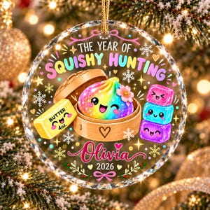 Könnte beinhalten: Ein runder, transparenter Acryl-Weihnachtsbaumschmuck mit dem Text THE YEAR OF SQUISHY HUNTING, Olivia und 2026. Das Motiv zeigt einen regenbogenfarbenen Teigkloß im Bambusdämpfer, ein gelbes Stück Butter mit Aufschrift 4oz und drei bunte Würfel.