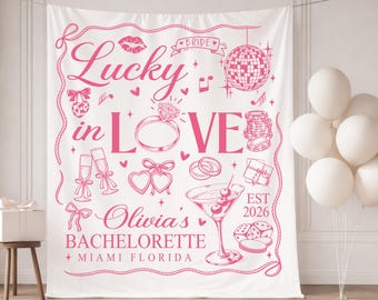 May include: A white bachelorette party backdrop with pink text and illustrations. The design includes the words "Lucky in Love," a diamond ring, champagne glasses, and a disco ball. The text "Olivia's Bachelorette + Miami Florida" is also featured.