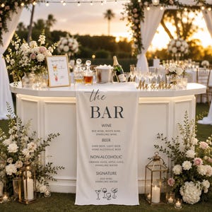 May include: A white bar decorated for a wedding, with a sign reading "The Bar". The sign lists drinks including wine, beer, and non-alcoholic options. Champagne, glasses, and floral arrangements are also present.