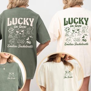 May include: Two oversized t-shirts, one olive green and one cream, feature the text "LUCKY in love" with decorative graphics. The olive shirt has the text "Emelias Bachelorette" and "Est 2025". The cream shirt has the same design. The bottom of each shirt has the text "The Bride".