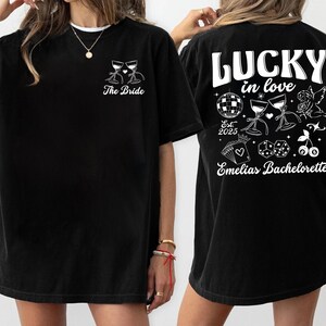 May include: Black t-shirt with white graphic designs. The front features two wine glasses and the text "The Bride". The back displays "LUCKY in love" with a disco ball, dice, cherries, and the text "Emelias Bachelorette".