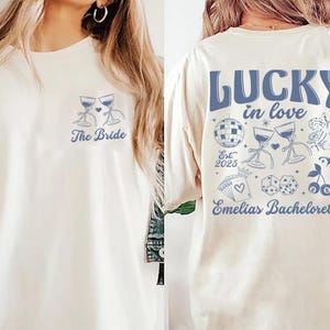 May include: Cream-colored t-shirt with blue graphics. The front features two wine glasses and the text "The Bride." The back displays "LUCKY in love" with additional illustrations and "Emelias Bachelorette" and "Est. 2025."