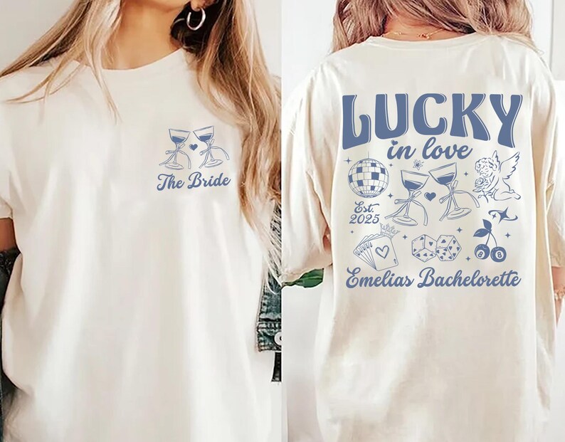 May include: Cream-colored t-shirt with blue graphics. The front features two wine glasses and the text "The Bride." The back displays "LUCKY in love" with additional illustrations and "Emelias Bachelorette" and "Est. 2025."