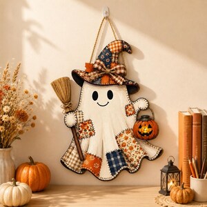 May include: A hanging wall decoration shaped like a ghost wearing a witch hat, featuring patchwork patterns in orange, blue, and white. The ghost holds a small broom and a jack-o'-lantern bucket, with stitched edges and a rope for hanging.