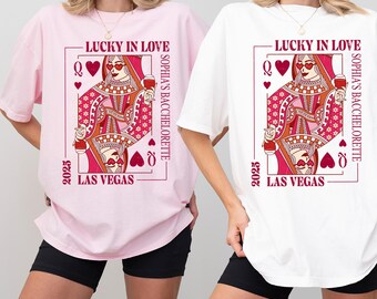May include: Two t-shirts, one pink and one white, each featuring a playing card design. The design includes the text "LUCKY IN LOVE," "SOPHIA'S BACHELORETTE," "2025," and "LAS VEGAS." The playing card depicts a queen holding a glass of wine.
