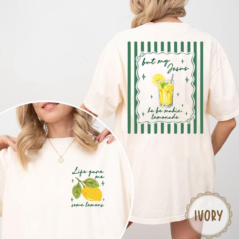May include: Ivory-colored t-shirt with a lemon and lemonade theme. The front features a lemon graphic with text, while the back displays a lemonade illustration with text. The design includes green accents and decorative elements.