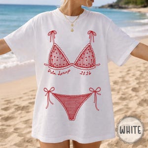 May include: White t-shirt with a red bikini design, including the text "Palm Springs 2026." The bikini top and bottom are outlined in red, with heart details. The shirt has a relaxed fit and short sleeves.