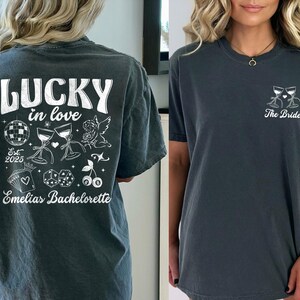 May include: Two dark gray t-shirts. One features the text "LUCKY in love" with illustrations of a disco ball, wine glasses, and dice. The other shirt has a wine glass design and the text "The Bride".