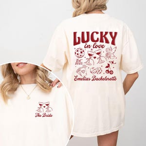 May include: Cream-colored t-shirt with burgundy text and graphics. The front features two wine glasses and the words "The Bride." The back displays "LUCKY in love" with additional illustrations and "Emelias Bachelorette" below.