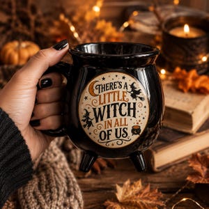 May include: A black ceramic cauldron-shaped mug with three small feet and a handle. The front features a circular cream-colored graphic with the text "There's a little witch in all of us," surrounded by illustrations of a crescent moon, stars, witches, and a cauldron.