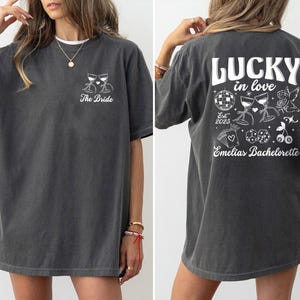 May include: Dark gray t-shirt with white graphic text and illustrations. The front of the shirt says "The Bride" with two wine glasses. The back says "LUCKY in love" with illustrations of a disco ball, wine glasses, and an angel.