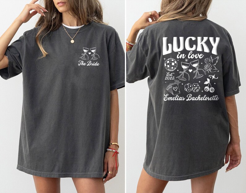 May include: Dark gray t-shirt with white graphic text and illustrations. The front of the shirt says "The Bride" with two wine glasses. The back says "LUCKY in love" with illustrations of a disco ball, wine glasses, and an angel.