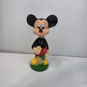 May include: A vintage Mickey Mouse figurine. The figure has a black body, large black ears, and a tan face. Mickey is wearing red shorts, yellow shoes, and holding a flower. The figurine is standing on a green base.