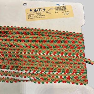 Peut inclure: Une bobine de tresse papillon décorative rouge, verte et dorée. La garniture est en viscose et polyester métallisé. L'étiquette indique "BUTTERFLY BRAID" et "MADE IN ENGLAND".