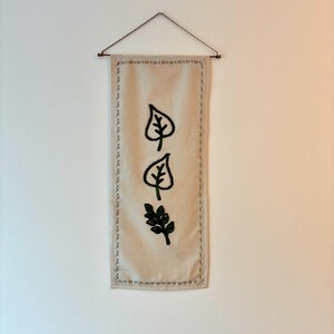 May include: A rectangular off-white fabric wall hanging featuring three embroidered leaf designs in dark green thread. The piece is suspended from a thin metal rod by a delicate copper-colored chain, with decorative stitching along the border.
