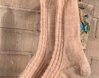 May include: A pair of light peach-colored knitted socks hanging on a clothesline. The socks are ribbed and have a textured pattern. They are clipped to the line with decorative clothespins against a brick wall.