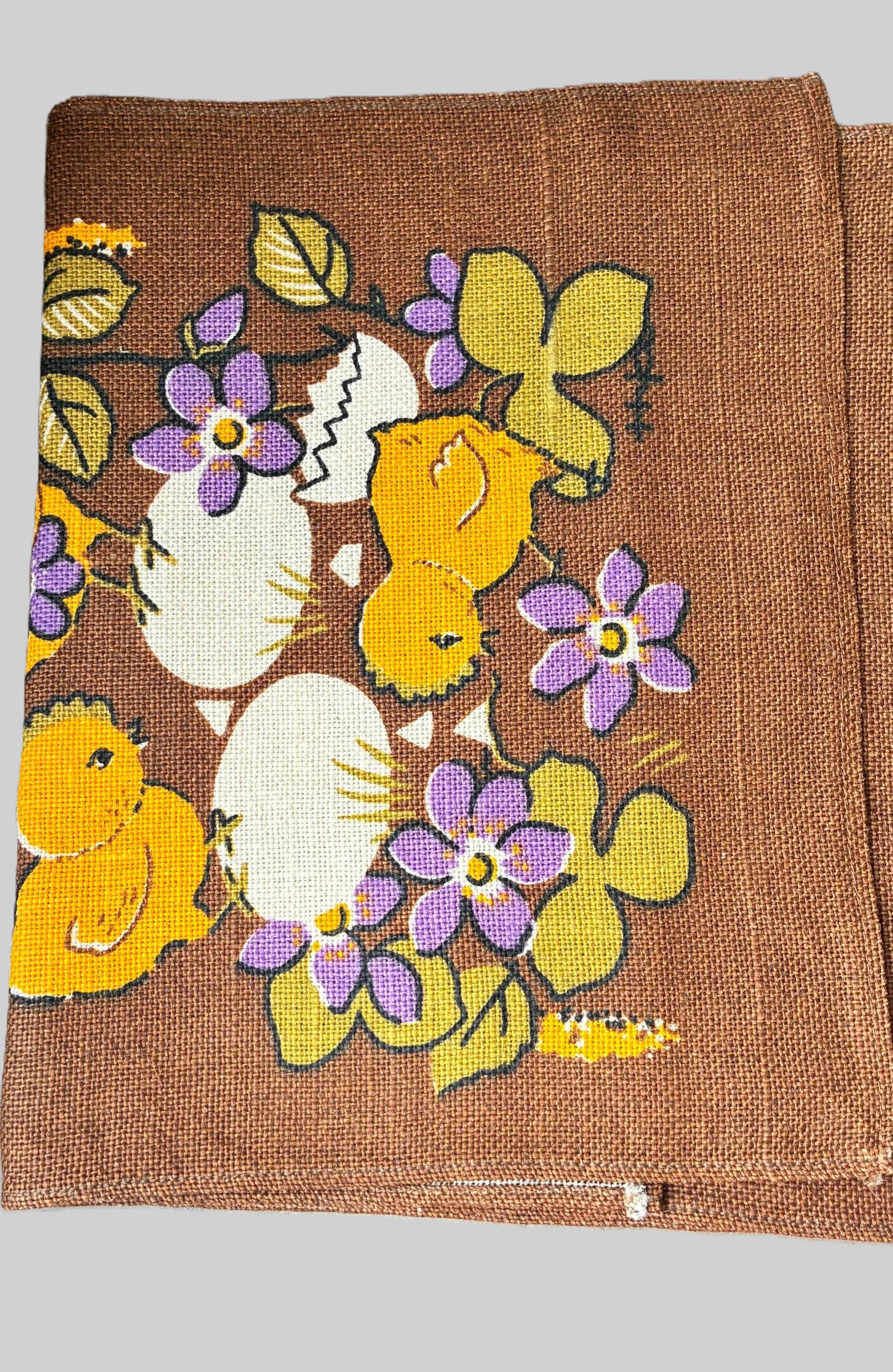 May include: Brown fabric featuring a repeating pattern of yellow chicks, white eggs, purple flowers, and green leaves. The design evokes spring and Easter. The fabric has a textured appearance.