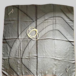 May include: A square scarf in a dark grey tone featuring a subtle snakeskin-like texture throughout. The design includes curved black lines and a central gold-toned buckle graphic. The word Guess is printed in white in two opposite corners.
