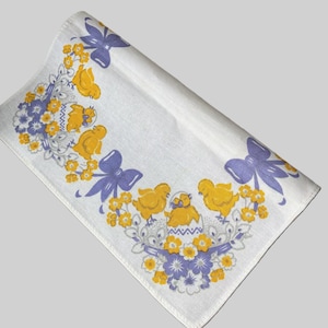 May include: White Easter-themed table runner with a repeating design of yellow chicks, purple bows, and floral accents. The runner is folded, showcasing the detailed print. A festive and decorative piece for the holiday.