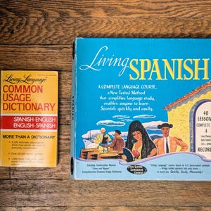 Op de afbeelding: Een blauwe doos met de tekst "Living Spanish" en een afbeelding van mensen in een Spaanse setting. De doos is gelabeld als een complete taalcursus met 40 lessen en 4 platen. Een geel en rood boek met de titel "Living Language Common Usage Dictionary" staat op de voorgrond.