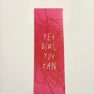 Puede incluir: Un marcapáginas rectangular con un fondo rosa vibrante que presenta un patrón caótico de finas líneas rojas garabateadas. El texto blanco impreso en el centro del marcapáginas dice YES GIRL YOU CAN, en un estilo de fuente simple, dibujado a mano y en mayúsculas.
