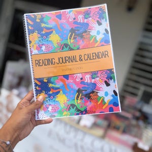 Puede incluir: Un diario de lectura y calendario de espiral con un patrón abstracto multicolor de salpicaduras de pintura y formas. Una banda horizontal naranja central muestra el texto READING JOURNAL & CALENDAR y HABONID GANDIA.