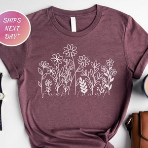 May include: A maroon t-shirt with a white floral design and the text "F*ck Trump". The shirt is styled with rolled-up sleeves and a knot at the waist. Accessories include a watch, earrings, and a coffee mug.