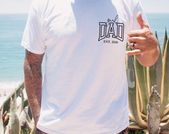 May include: White t-shirt with the text "girl DAD EST. 2026" in black. The person is wearing brown camouflage shorts. The background includes a beach, ocean, and green plants.