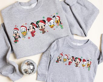 May include: Two gray sweatshirts, one adult and one child size, featuring a festive design. The design includes cartoon characters like Mickey Mouse, Donald Duck, and Goofy, all wearing Santa hats and scarves. A small disco ball is in the image.