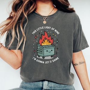 May include: A gray t-shirt with a graphic of a burning trash can with a smiling face and the text "This little light of mine, I'm gonna let it shine."