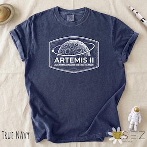 May include: A navy blue t-shirt with a white graphic of a planet with the text "ARTEMIS II 2026 Manned Mission Orbiting the Moon." The shirt also has the words "TRUE NAVY" printed on the bottom left.