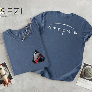 May include: Two blue t-shirts, one folded with a silver chain necklace and a patch featuring a rocket and moon. The other shirt has "ARTEMIS II" printed on the back. A toy rocket and a lunar map are also visible.