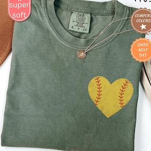 Puede incluir: Una camiseta verde con un gr&aacute;fico de pelota de softball en forma de coraz&oacute;n amarillo. La camiseta tiene un aspecto desgastado y est&aacute; hecha de una tela suave y c&oacute;moda. El texto "COMFORT COLORS" est&aacute; impreso en la etiqueta.