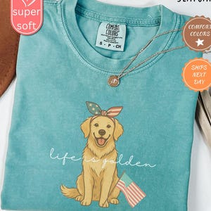 May include: A teal blue t-shirt with a golden retriever wearing a red, white, and blue bandana and holding a small American flag. The text "life is golden" is printed on the shirt.