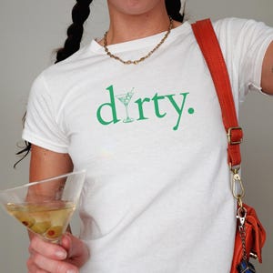 May include: White t-shirt with the word "dirty" in green, featuring a martini glass graphic. The shirt is paired with a red shoulder bag and a gold chain necklace. A martini glass with olives is held in the hand.