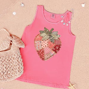May include: Pink tank top with a large strawberry applique made of various patterned fabrics. A woven beige handbag is on the left. A beaded necklace is draped on the shoulder. The background is a sandy beach.