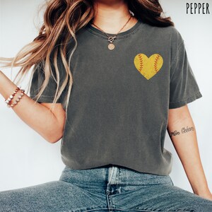 Puede incluir: Una camiseta gris con un gr&aacute;fico de softball en forma de coraz&oacute;n amarillo. La pelota de softball tiene costuras rojas.