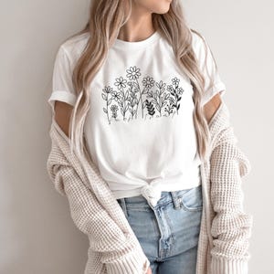 May include: White t-shirt featuring a black floral line art design. The design includes various flowers and foliage, with the word "flowers" written below. The shirt is paired with blue jeans and a cream-colored cardigan.