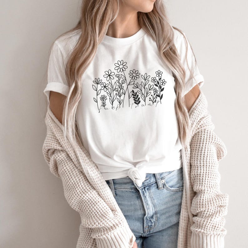 May include: White t-shirt featuring a black floral line art design. The design includes various flowers and foliage, with the word "flowers" written below. The shirt is paired with blue jeans and a cream-colored cardigan.