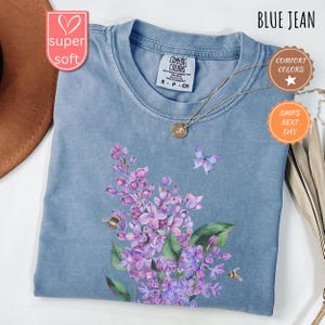 May include: A blue jean coloured t-shirt with a floral design featuring purple flowers, green leaves, a butterfly, and bees. The shirt has a crew neck and short sleeves. The text "BLUE JEAN" is at the top right.