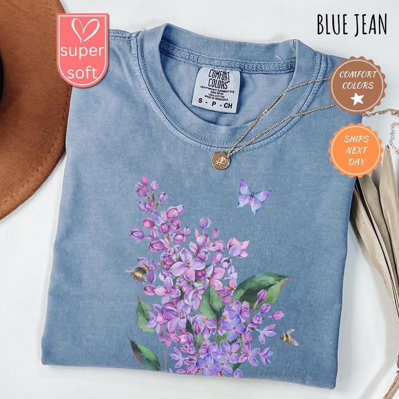 May include: A blue jean coloured t-shirt with a floral design featuring purple flowers, green leaves, a butterfly, and bees. The shirt has a crew neck and short sleeves. The text "BLUE JEAN" is at the top right.