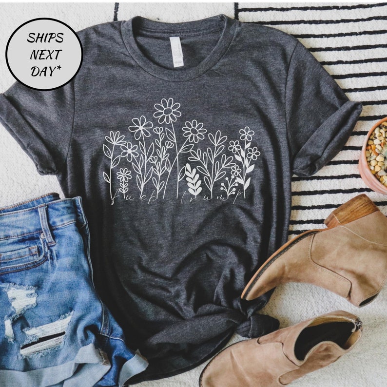 May include: Dark gray t-shirt with a white floral design and the text "fuck trump". The shirt is paired with denim shorts and tan boots. The image is on a striped rug.