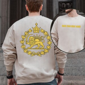 May include: Beige sweatshirt featuring a yellow emblem of a lion holding a sword, a crown, and a wreath. The back of the sweatshirt has the text "MAKE THE GREAT AGAIN" in yellow. The sweatshirt is made of a soft material.