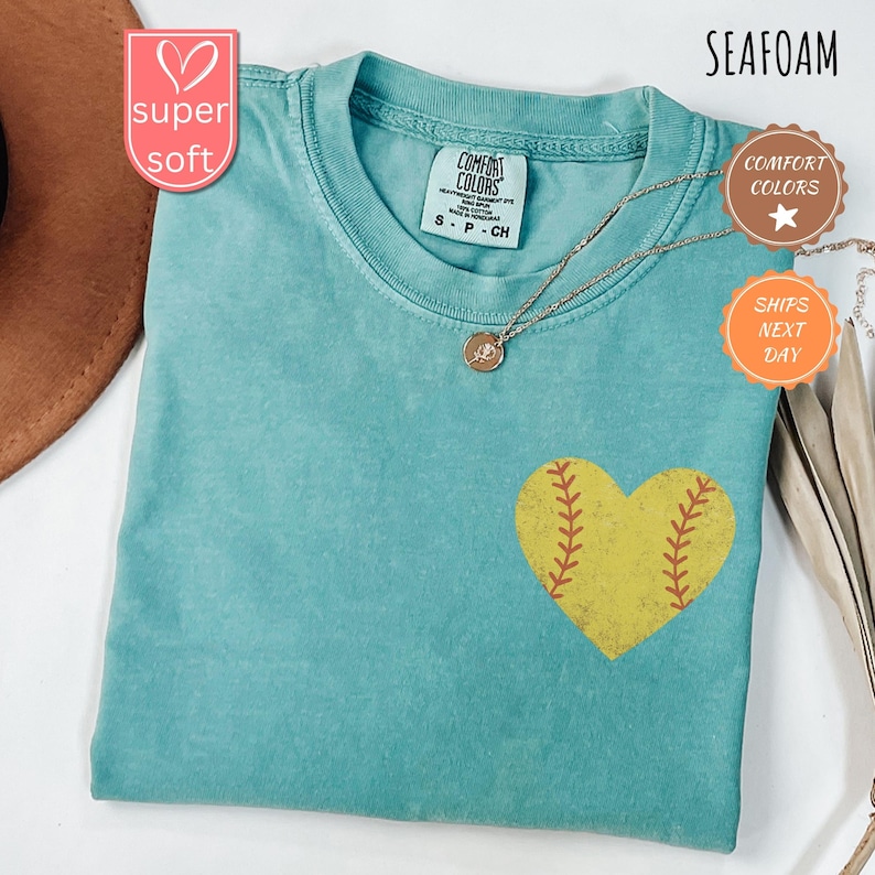 Puede incluir: Una camiseta azul turquesa con un gr&aacute;fico de softball en forma de coraz&oacute;n amarillo. La camiseta est&aacute; hecha de una tela suave y c&oacute;moda y tiene un aspecto desgastado. El texto "COMFORT COLORS" est&aacute; impreso en la etiqueta.